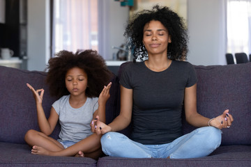 Mindful african mom with cute funny kid daughter doing yoga exercise at home, calm black mother and mixed race little girl sitting in lotus pose on couch together, mum teaching child to meditate