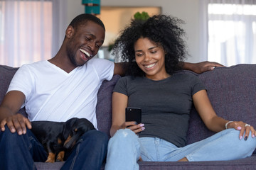 Happy black african couple with pet laughing looking at smartphone taking selfie, having fun in...
