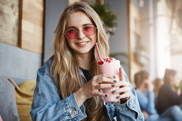 Take pause and drink tasty cocktail. Outdoor shot of carefree cute blond female student in stylish pink sunglasses, holding drink with strawberry and gazing with caring smile at camera