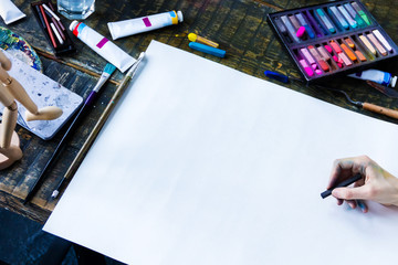 Soiled artists hands is painting on blank paper sheet. Top view of creative workplace on old wooden table with different painting tools, brushes, colorful paints, pastels, tubes. Art concept.