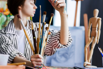 Woman artist at her creative workplace holds bunch of used wooden paintbrushes in soiled with paints hands. Workshop with painting tools and paintings on easels on background. Art concept.