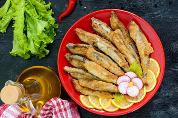 Fried capelin with lemon on a red plate on a black wooden background. A dish of small sea fish. Top view. Flat lay
