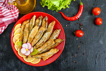 Fried capelin with lemon on a red plate on a black wooden background. A dish of small sea fish. Top view. Flat lay