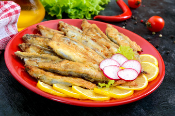 Fried capelin with lemon on a red plate on a black wooden background. A dish of small sea fish.