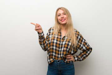 Blonde young girl over white wall pointing finger to the side and presenting a product