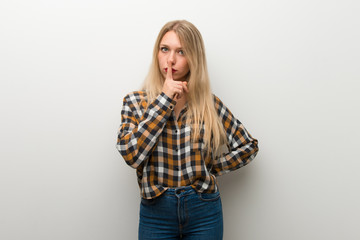 Blonde young girl over white wall showing a sign of silence gesture putting finger in mouth