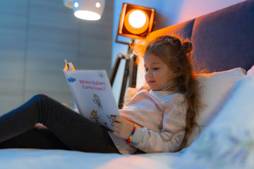 Small pretty long-haired girl in a sweater laying in bed with the book