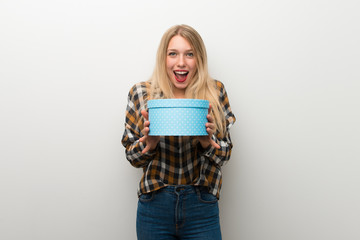 Blonde young girl over white wall surprised because has been given a gift