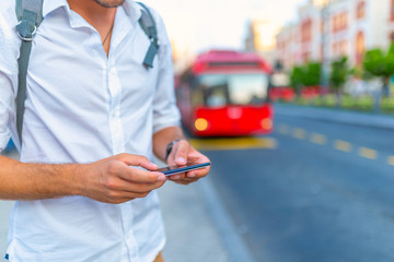 Young man with luggage bag walking in the city street looking at smartphone, using app, gps,...