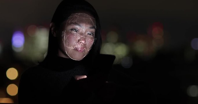 Asian Woman at night using facial recognition technology to unlock smart phone on street
