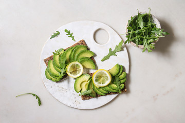 Vegan sandwiches with sliced avocado and lemon on rye bread, arugula salad served on ceramic board over white marble background. Flat lay, space