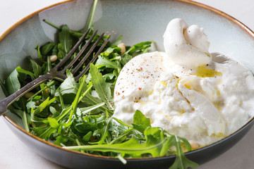 Sliced Italian burrata cheese, fresh arugula salad, pine nuts and olive oil in white ceramic plate...