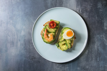 Vegetarian sandwich with sliced avocado, sun dried tomatoes, egg, shrimps, served on ceramic plate over blue texture background. Flat lay, space