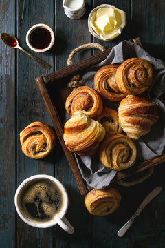 Variety of homemade puff pastry buns cinnamon rolls and croissant served with coffee cup, jam, butter as breakfast over dark plank wooden background. Flat lay, space