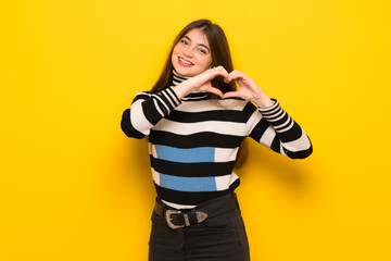 Young woman over yellow wall making heart symbol by hands