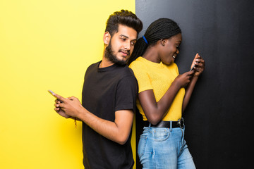 Young happy mixed race couple, indian man spy on african woman use phone standing back to back on the middle on different yellow and black background