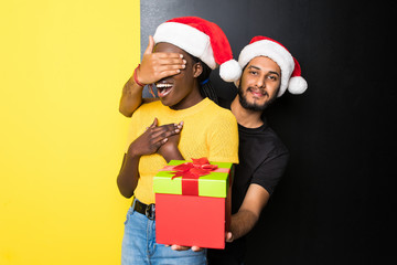 Young happy interracial couple, indian man make a surprise with Christmas gift for his african woman wearing santa hats standing isolated on yellow black background