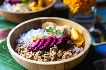 Close up smoothie bowl made with mango, banana, granola, grated coconut, dragon fruit, chia seeds and mint on colored background.