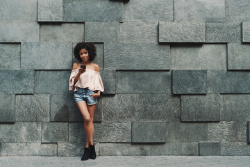 A beautiful cheerful African-American woman with a medium curly Afro hair is leaning against the wall on stony blocks and checking her phone with a copy space area on the right for your ad text - Powered by Adobe