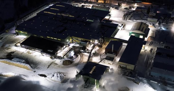 4K aerial hyperlapse of a busy wood factory at winter night. Tractors feeding timber to the production building.