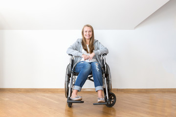 Happy invalid woman is sitting on a disability chair in a white empty room.