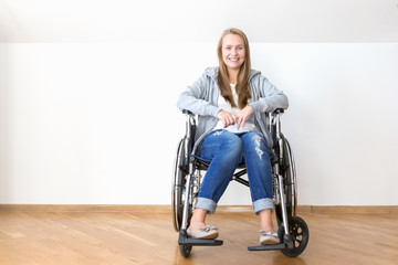 Happy invalid woman is sitting on a disability chair in a white empty room.