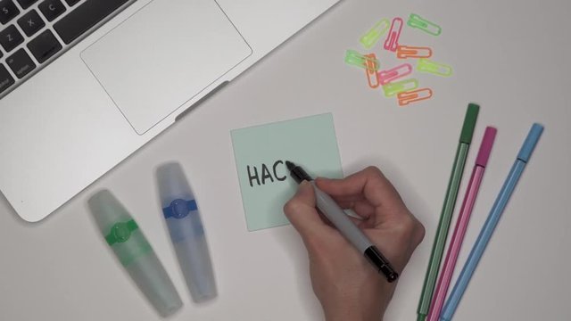 Woman writing HAKING on adhesive notepad. Laptop and stationery on the table.