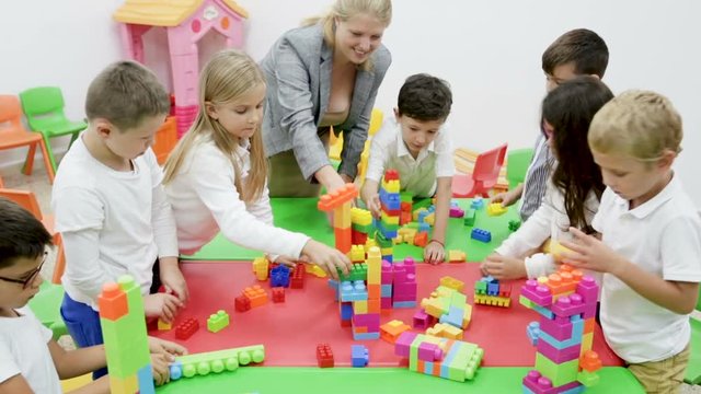 Group of schoolkids playing with toy bricks in circle with their cheerful female teacher 