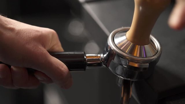 A close-up of a hand holding a portafilter with ground coffee on the edge of the table, another hand presses it with a tamper. Coffee making in a bar
