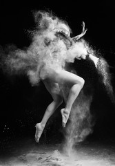 Schönes schlankes Mädchen, das einen gymnastischen Bodysuit trägt, der mit Wolken des fliegenden weißen Pulvers bedeckt ist, springt tanzend auf eine Dunkelheit. Künstlerisches konzeptionelles und werbliches Schwarz-Weiß-Foto.
