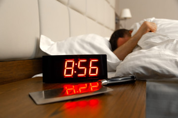 Alarm clock on bedside table with red numbers, kitchen knife, sleeping man in bed in room. Concept...
