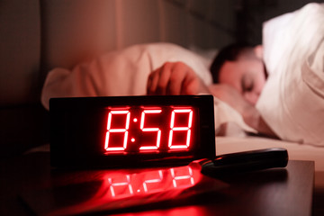 Alarm clock on bedside table with red numbers, kitchen knife, sleeping man in bed in dark room....