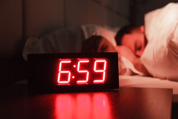 Rectangular alarm clock on the bedside table with red numbers, sleeping man in bed in dark room. Concept chef sleeping after hard day at restaurant, sleep disturbance, oversleep work, night shift - obrazy, fototapety, plakaty