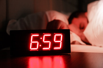Rectangular alarm clock on the bedside table with red numbers, sleeping man in bed in dark room....