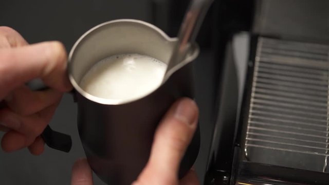 A close-up of hands holding a jug with milk and a steam wand heating it at the coffee machine. Coffee making in a bar
