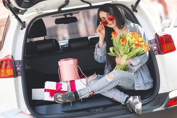 Young attractive woman in the car with presents box, gift and flowers. Beautiful lady in spring time with bouquet of tulips. Female in the automobile. © olenachukhil