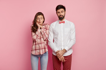 Serious handsome young man with thick stubble, keeps hands together, wears elegant white shirt, bowtie and red trousers, feels shy in presence of beautiful woman, puzzled during first date with lover