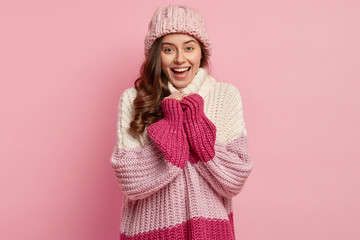 Studio shot of joyful pretty woman wears loose knitted pullover and hat, being in good mood after walk on frosty winter day, smiles broadly, isolated over pink concept. People, season concept