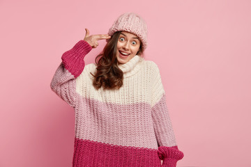 Horizontal shot of good looking woman makes suicide gesture, smiles positively, pretends killing herself and shooting in temple, dressed in oversized warm winter jumper, hat, isolated on pink wall