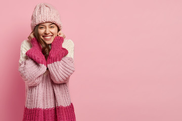 Happy attractive female youngster keeps hand near ears, dressed in warm knitted sweater and...