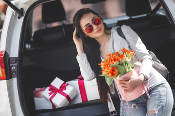 Young attractive woman in the car with presents box, gift and flowers. Beautiful lady in spring time with bouquet of tulips. Female in the automobile. © olenachukhil