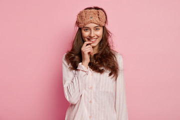 Healthy sleep concept. Satisfied young woman with pleasant smile, has sleep mask on head, dressed in pyjamas, feels refreshed after having good rest and seeing dreams, isolated on pink wall.