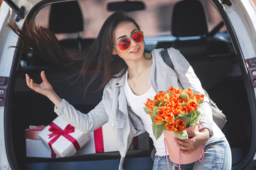 Young attractive woman in the car with presents box, gift and flowers. Beautiful lady in spring time with bouquet of tulips. Female in the automobile. © olenachukhil