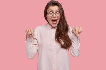 Pleased pretty European woman points down with both index fingers, wears optical glasses, dressed in stylish shirt, isolated over pink background. People, promotion and good emotions concept © WHstudio Leushin N