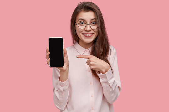 Shot of good looking woman with satisfied expression, points at modern electronic device, shows empty space on display, smiles gladfully, wears fashionable shirt, isolated over pink background