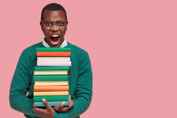 Photo of annoyed black young man screams with irritation, holds pile of books, dressed in casual...