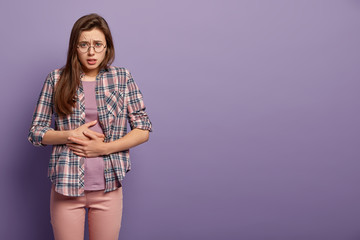 Painful feelings concept. Upset woman feels hungry, keeps hands on belly, suffers from pain, wears...