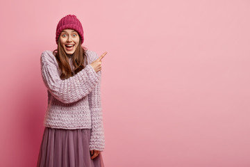 Studio shot of pleased woman with positive smile, points with fore finger, wears knitted sweater...