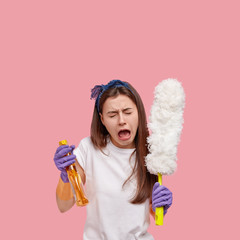 Desperate mad housemaid cries from despair, feels overworked after cleaning hotel room, wears purple rubber gloves, casual t shirt, isolated over pink background empty space above for your information