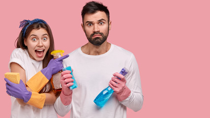 Fototapeta premium Happy young woman with cheerful expression, feels surprised, stands near bearded guy, hold detergents and sponges, isolated over pink background with free space for your promotional content.